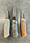 Tre ferdige knivar med mønstersmidde blad. Kniven til venstre har skaft av kvalkjeve, den i midten er av valbjørk, medan skaftet på den høgre er av bjørkeknute.