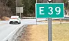 Vegskilt for E39 ved ein svingete veg med bil i bakgrunnen.