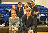 Seks unge personar står og sit samla framfor blå stolar i eit auditorium.