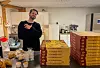 Mann ved disk med store stablar pizzaesker og tilbehør i eit kjøkkenrom.